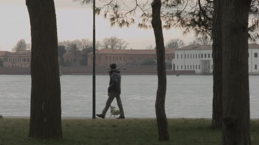 A man walking a dog along a river edge with some buildings in the background