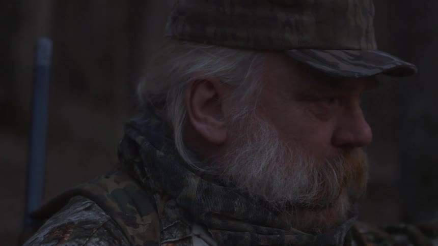 An older man with long hair and a beard wearing hunting gear outside at night