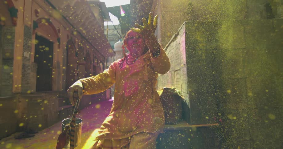 A woman sits on a ledge with her hand raised and colored powder falls onto her from above