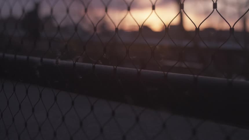 A sunset and buildings are seen from behind a chain-link fence
