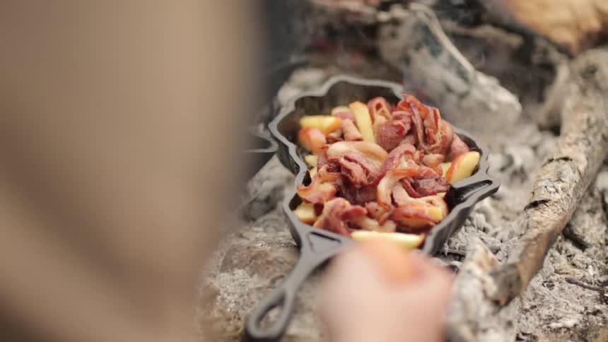 A person cooking bacon, potatoes and eggs in an iron skillet over a campfire
