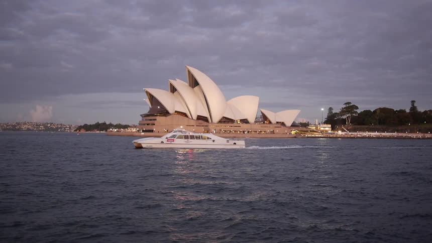 A boat sailing through the water near a large building at shore on a grey and cloudy day