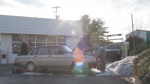 A man looks under the hood of a car parked on the side of a convenience store