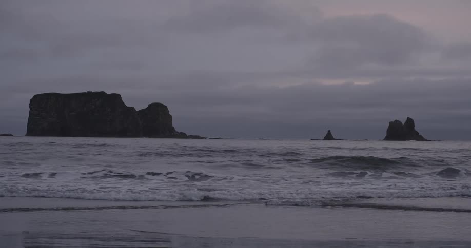 A beach on a cloudy day with rocky islands in the distance and waves rippling toward the shore