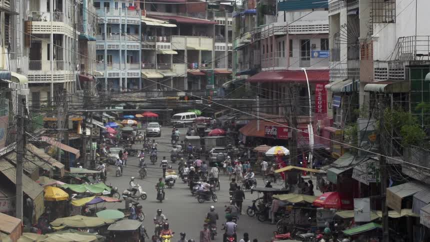 Numerous people riding scooters on a busy city street surrounded by tall buildings and vendor tents