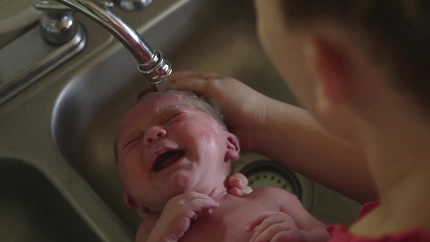 An infant is bathed under a kitchen faucet
