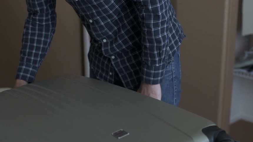 A man leans over and picks up a suitcase from a closet and places it on a bed before opening it