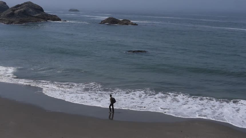 A man wearing a backpack walking along the beach as waves roll in