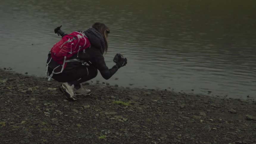 A woman wearing a pink backpack squatting in front of a body of water while looking at a camera