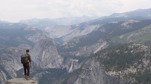 A man sits on a high rock overlooking a valley