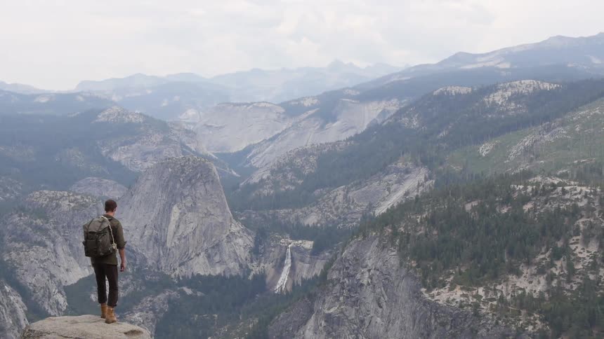 A man sits on a high rock overlooking a valley