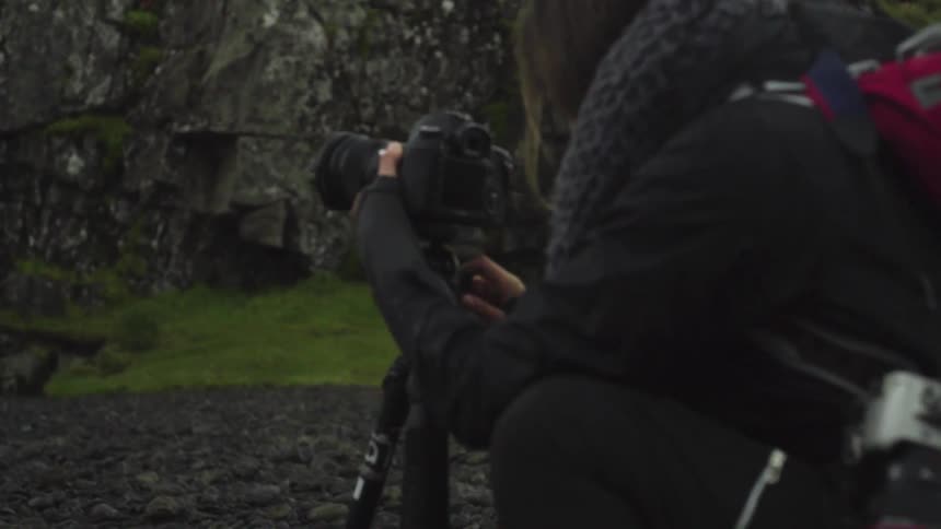 A woman wearing a backpack films a waterfall in the mountains at sunset
