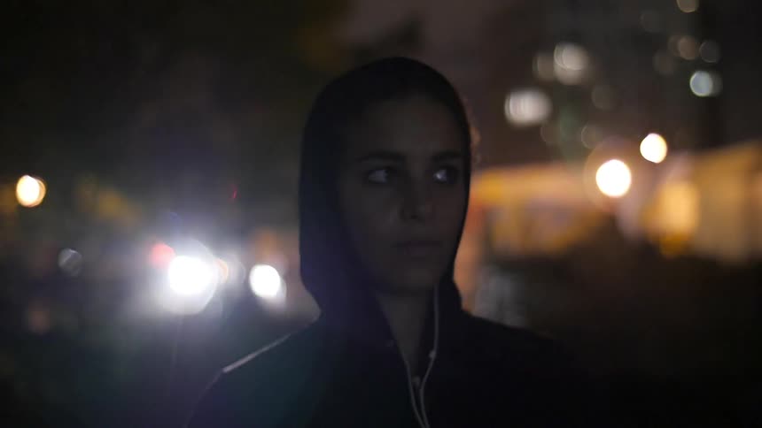 A woman stands in the city at night listening to music on her headphones and nodding her head