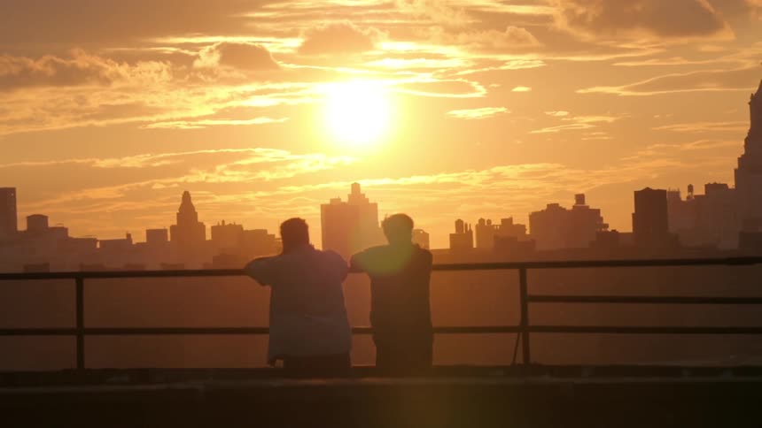 Two people lean against a railing while watching a brilliant sunset behind a city skyline