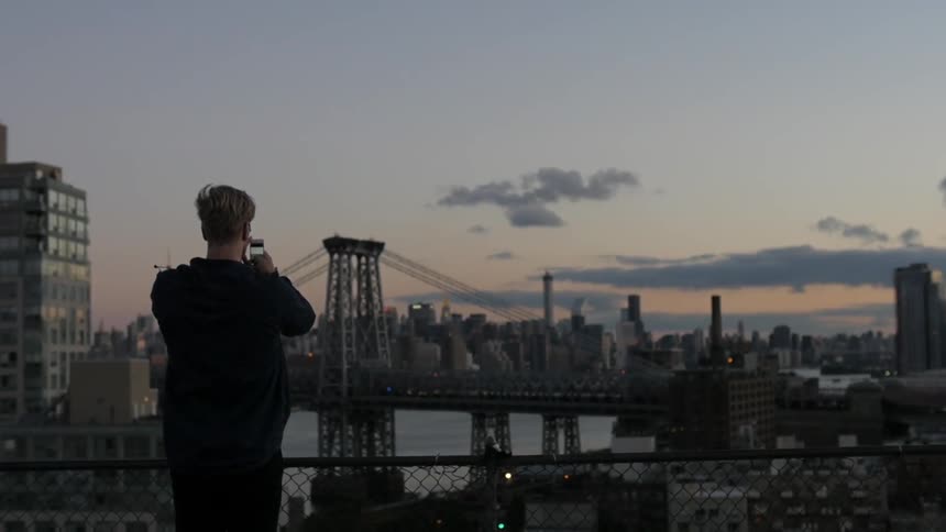 A man stands and photographs a bridge and a city with his phone at sunset