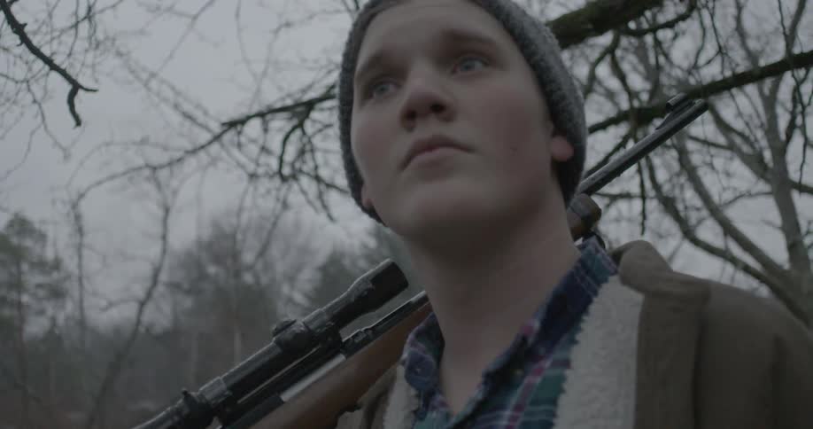 A young man stands holding a gun over his shoulder in the woods