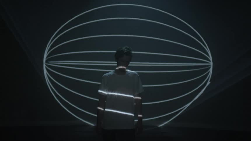 A man stands facing away from a big screen as different lights are projected onto the screen and himself