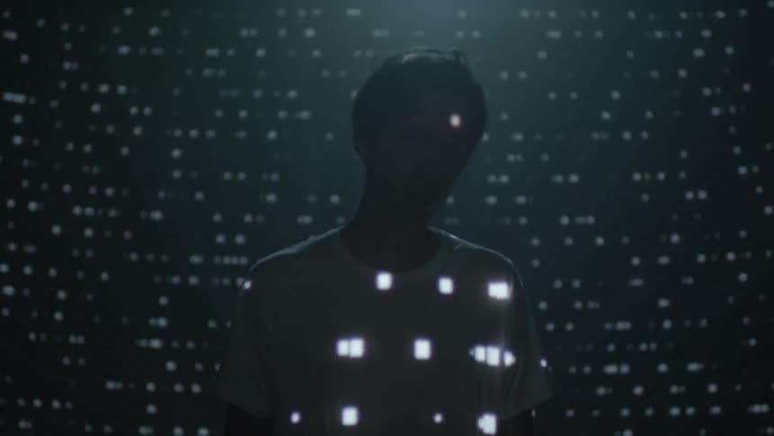 A young man stands in a dark room while animated light patterns are projected onto him and the wall behind him