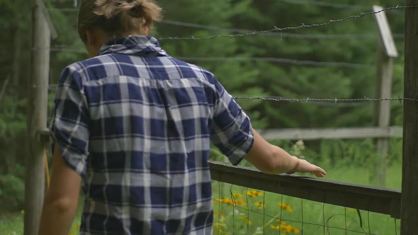 A young woman walks next to a fenced-in, overgrown garden while touching the fencing and looking down at the ground