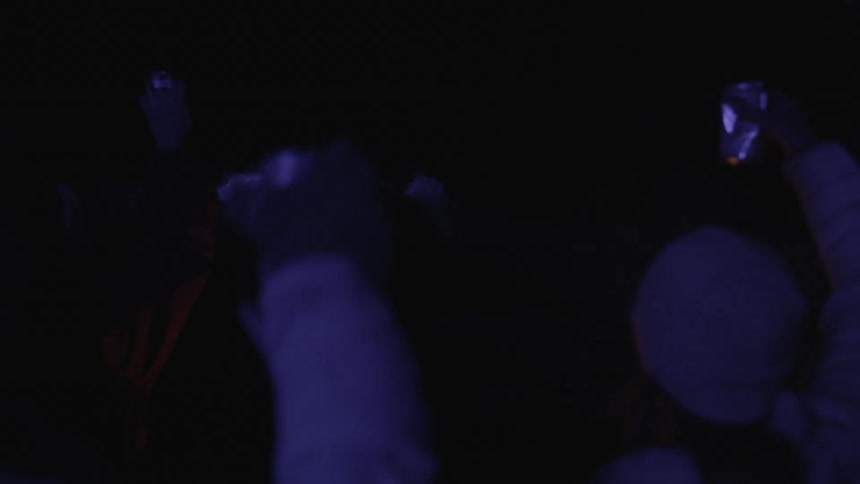 A crowd of people waving their hands in the air holding cans at night while a person kayaks in the water