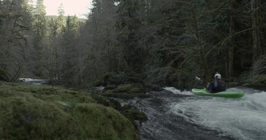 A man kayaks through rapids while another backflips from a ledge